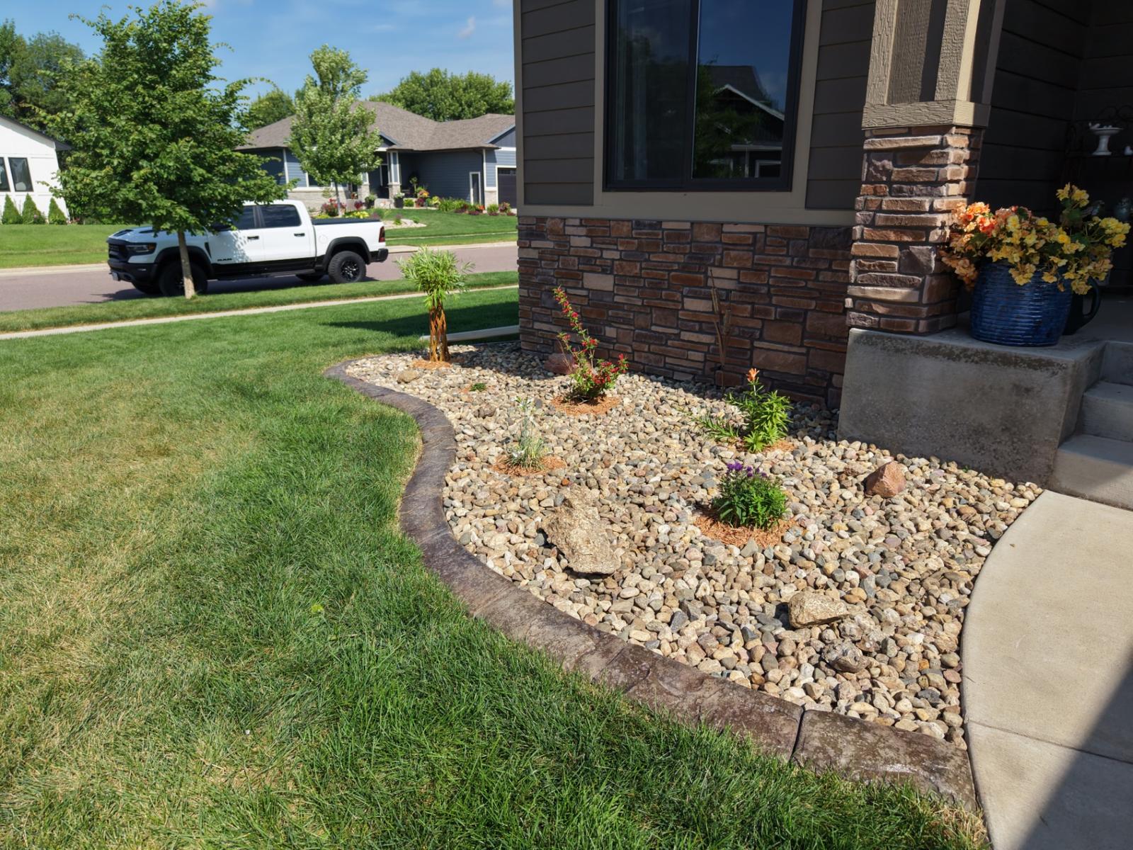 After — front entry rock bed with curved stamped edging, fresh plantings, and accent boulders against the stone facade