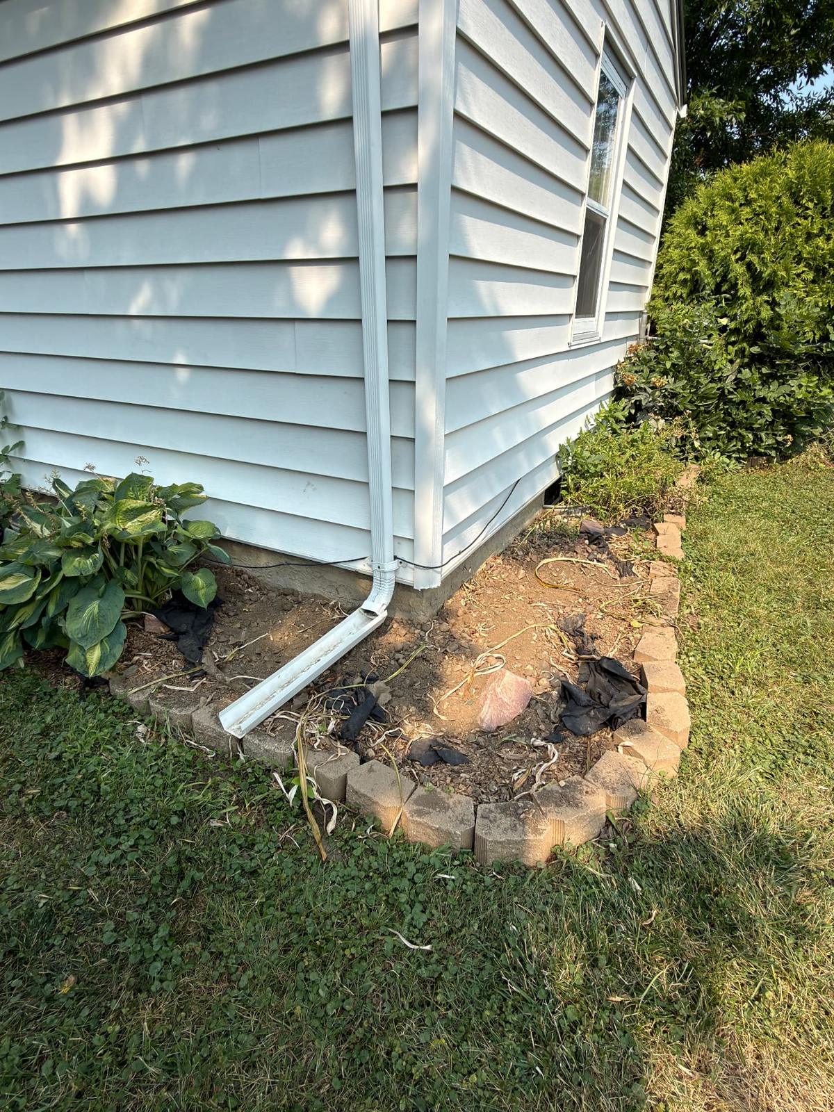 Before — corner bed with failed fabric, old block edging, and a downspout dumping water right against the foundation