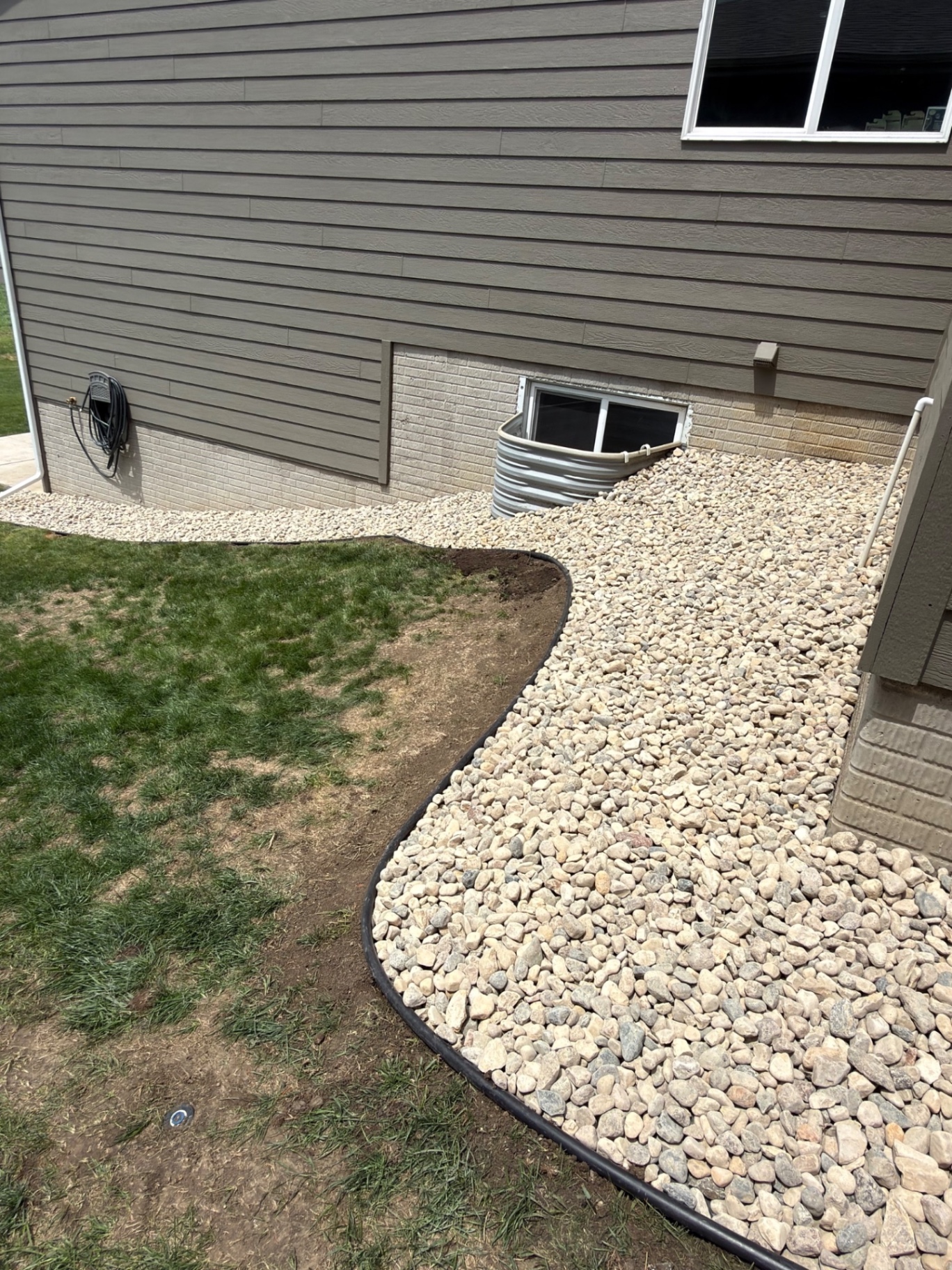 After — finished river rock bed wrapping around the window well on the side of the house with smooth curved edging