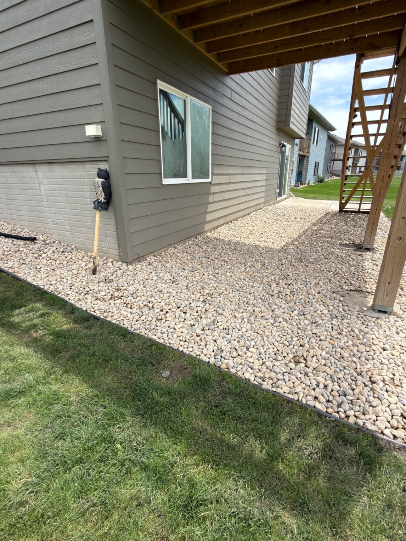 After — wide river rock bed wrapped around the back corner of the house under the elevated deck with sharp edging against the lawn