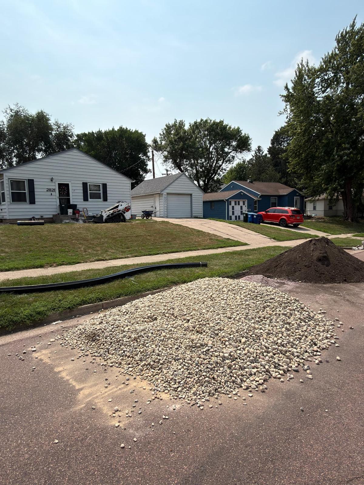 During — Bobcat mini skid steer on site with piles of new river rock, black soil, and plastic edging staged for the install