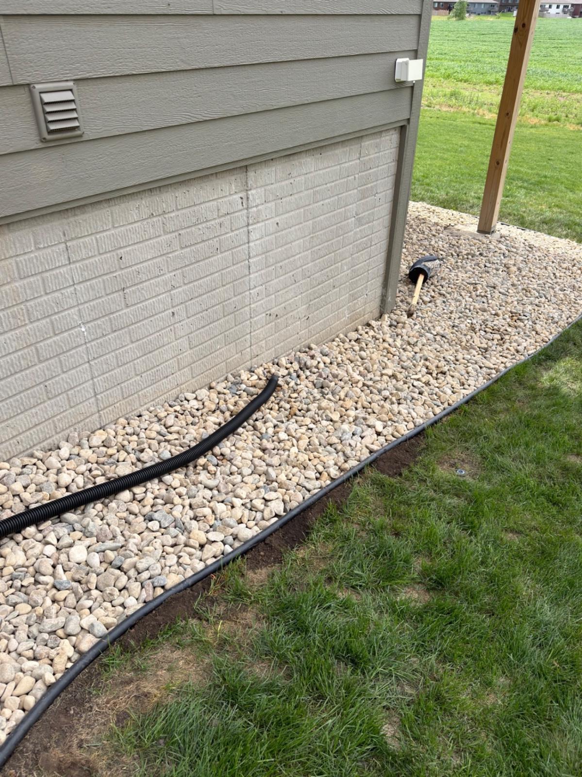 After — finished river rock bed along the foundation with french drain line and clean steel edging against the grass