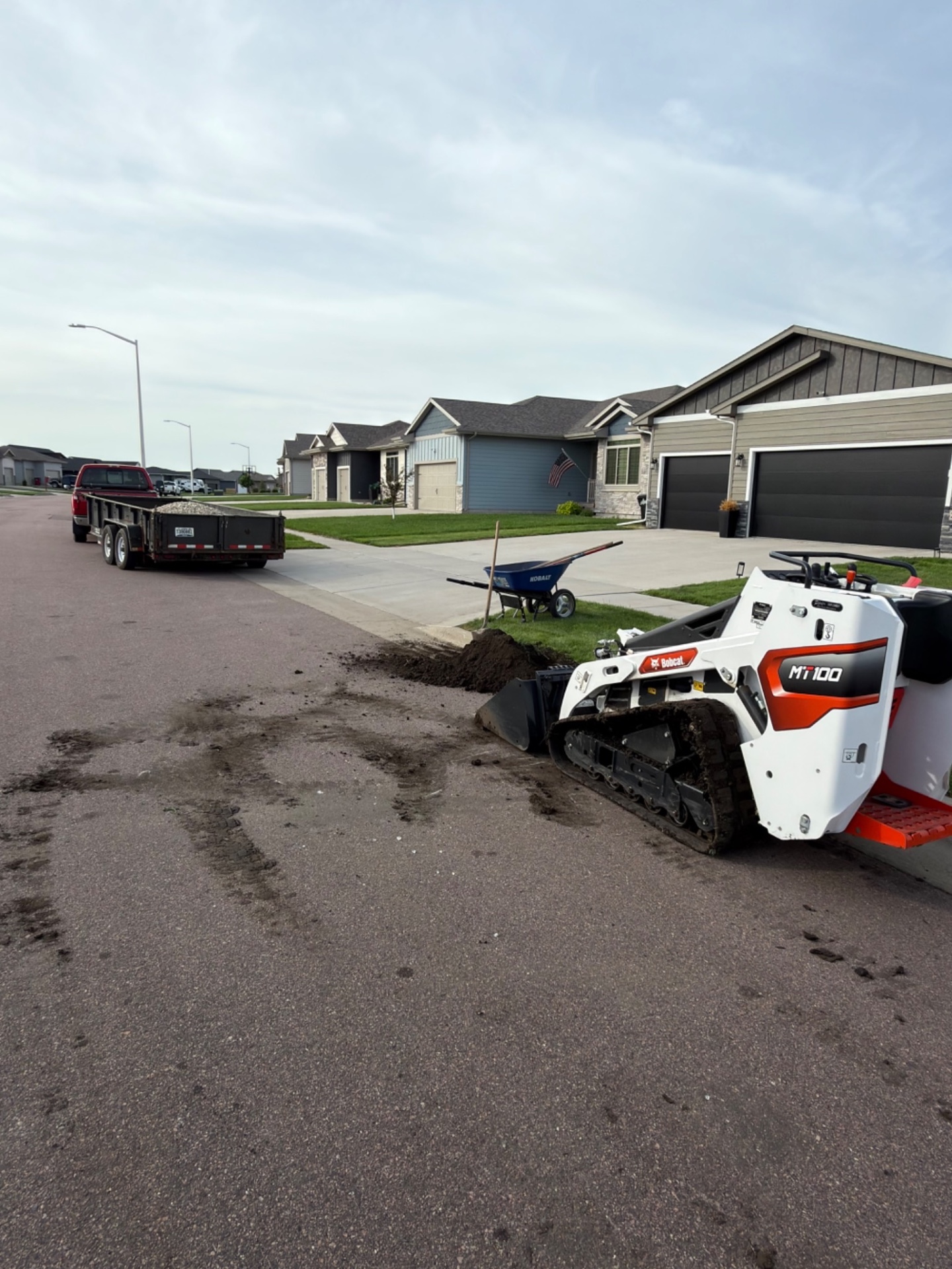 During — Bobcat MT100 mini skid steer staged at the curb with dump trailer of rock parked on the street