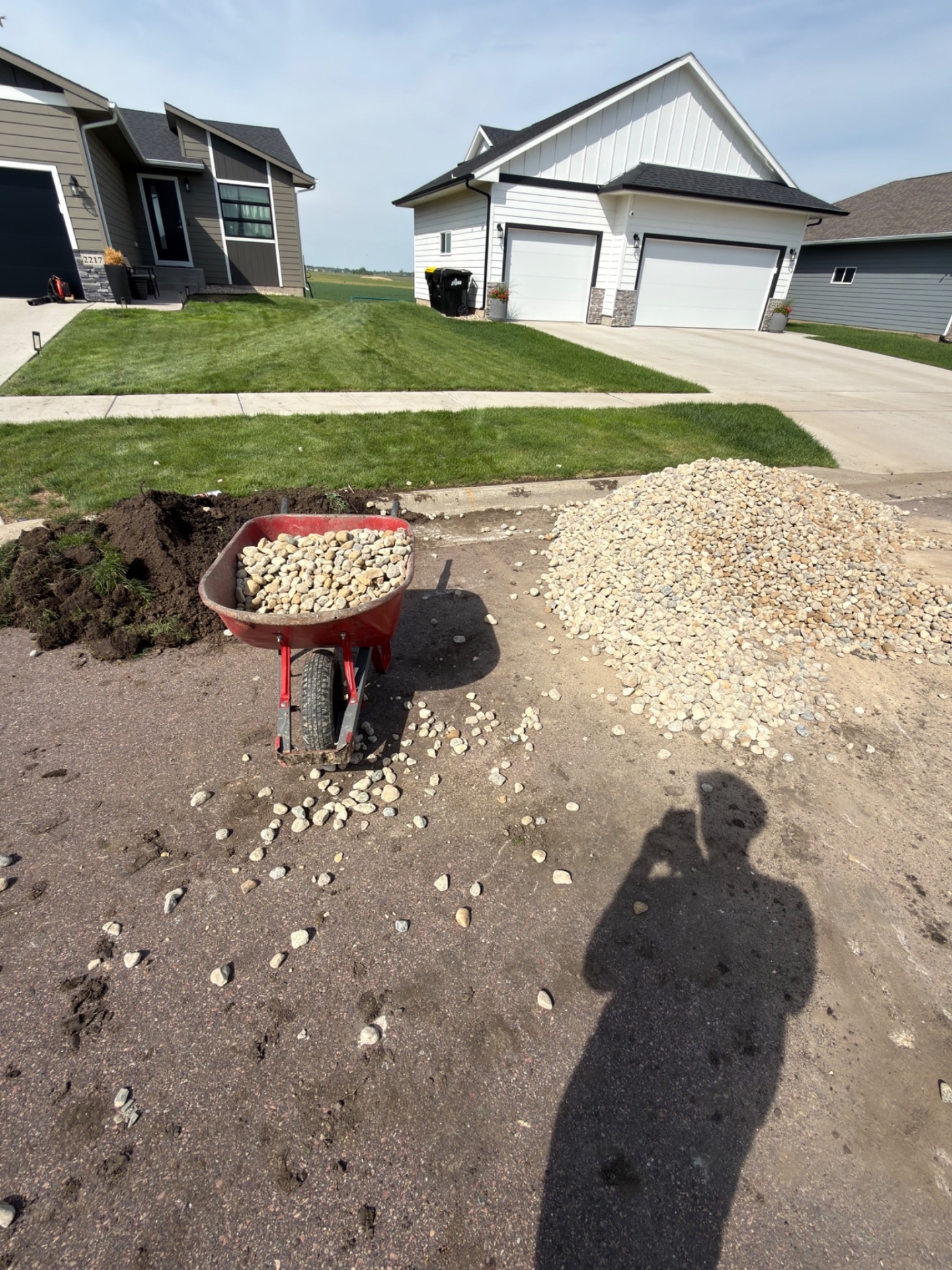 During — wheelbarrow loaded with rock between a pile of dirt and a fresh pile of river rock on the driveway