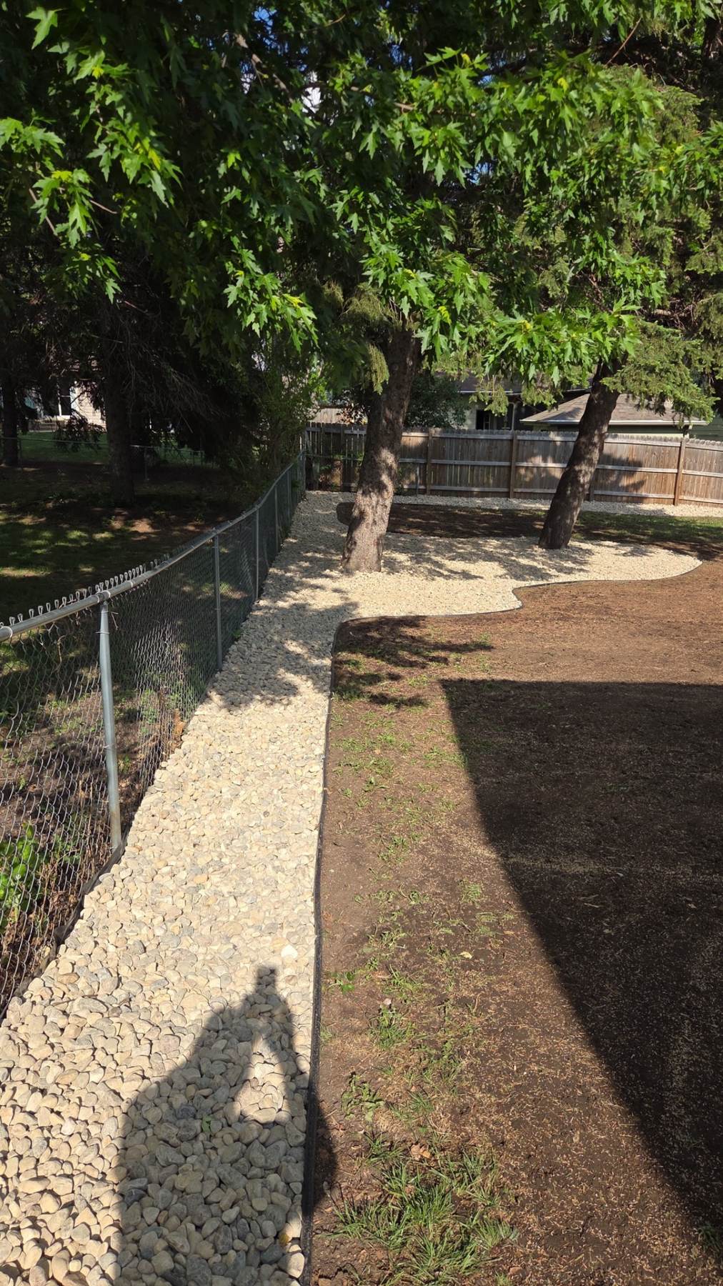 After — long rock drainage strip running along the chain link fence with fresh tree beds